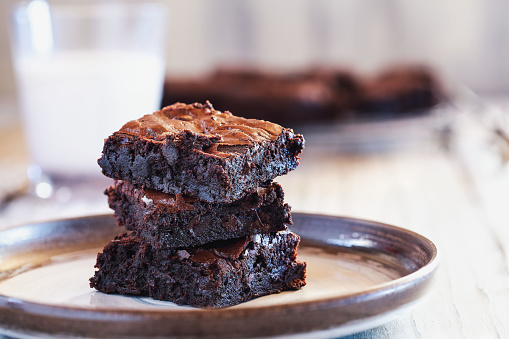 Fresh made homemade fudgy brownies stacked on a saucer over a white rustic wooden table. Extreme shallow depth of field with blurred background and a glass of milk.