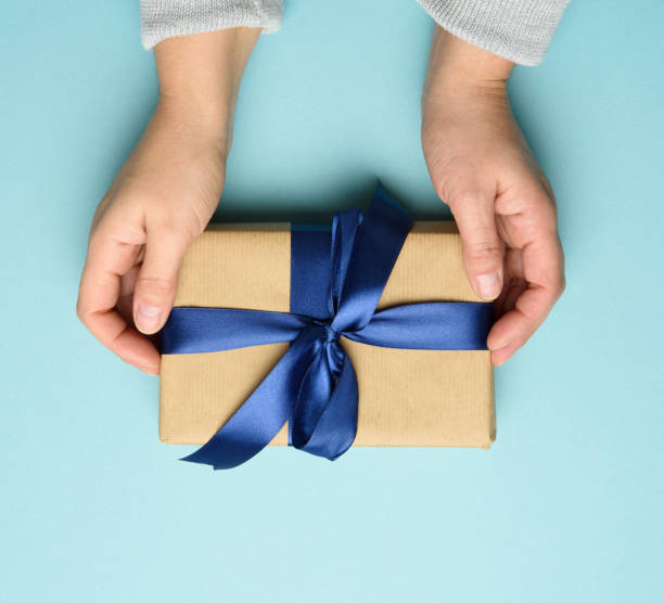 istock two female hands in a sweater hold a square gift box with a blue bow on a blue background 1289902401