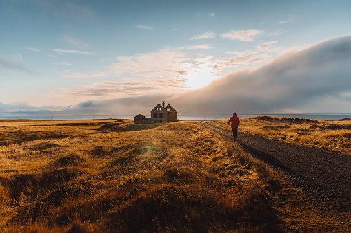 Man traveler walking at the beautiful golden meadow near the ocean to the old house during sunny autumn sunset on Snaefellsnes Peninsula - West Iceland
