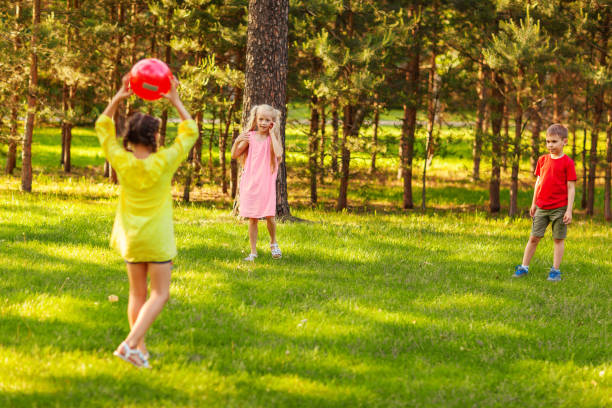 duas meninas e meninos jogando bola vermelha no gramado verde no parque no dia de verão - playing the ball - fotografias e filmes do acervo