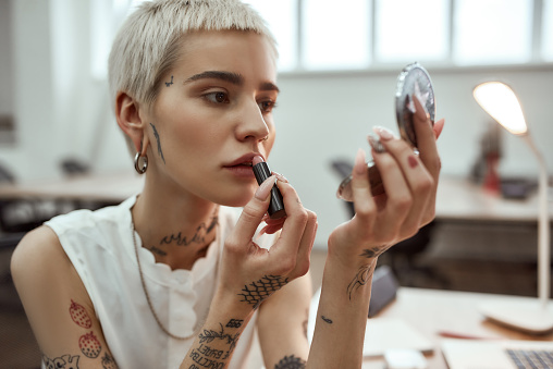 Portrait young and attractive blonde tattooed woman applying lipstick while sitting in the modern office. Doing make up. Business concept. Coworking space