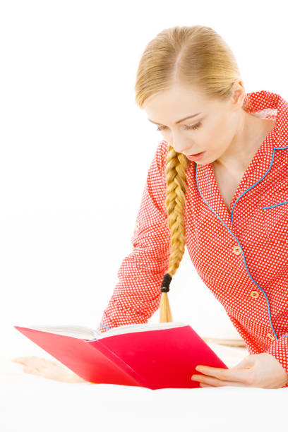 Girl lying in bed reading book. Young blonde female wearing red dotted pajamas relaxing at home on mattress.