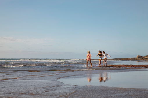 El Medano, Tenerife, Canary Islands, Spain - August 21, 2020: group of three young women wearing swimsuits or casual summer clothes, walking barefoot on the beach, while talking, laughing and enjoying.