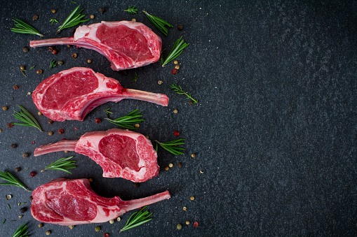 Raw Lamb Chops on a dark slate chopping board, prepared with fresh herbs, peppercorns and rock salt. Overhead view with copy space. Stock photo.