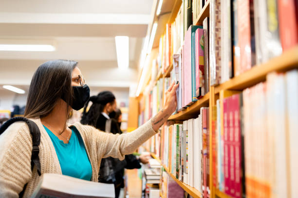 istock Student at a library choosing a book in the shelf, using face mask 1289937024