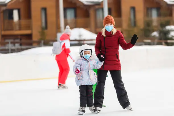 young mother teaching her little daughter ice skating at outdoor skating rink. Family wearing a medical mask during COVID-19 coronavirus enjoy winter on ice-rink outdoors