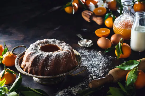 Bundt cake made of tangerine clementines baked homemade with ingredients on moody dark background