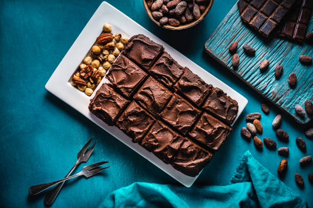 Brownie chocolate cake on turquoise background with nuts forks and linen napkin, cocoa beans and rustic chocolate bar