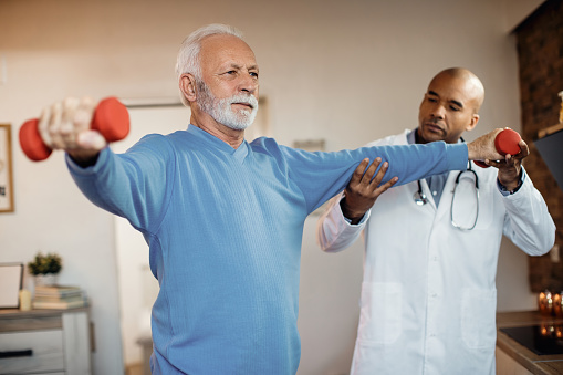 Senior man having physical therapy with African American doctor and exercising with dumbbells at nursing home.