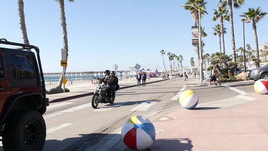 Oceanside, California USA - 8 Feb 2020: People walking, waterfront promenade near pier, pacific ocean tropical beach tourist resort and palm trees. Couple on motorcycle, beachfront street on sunny day