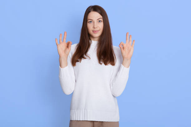 istock Young beautiful confident female showing ok sign with both hands, looks at camera, posing isolated over blue background, wearing white sweater, having brown straight hair. 1289951019