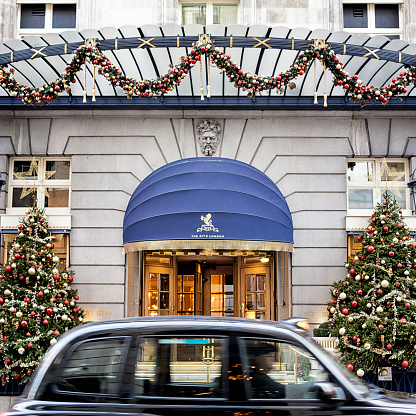 London, UK - December 5, 2020: A black cab driving outside the main entrance of The Ritz Hotel Piccadilly in London at day. The hotel is beautifully ornamented with elegant Christmas decoration for the festive season. The Ritz, opened in 1906, is a luxury hotel and international landmark in London's West End. (Panoramic composite of two images)