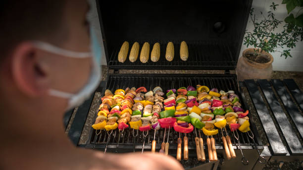 Chef using a face mask and cooking a skewers with corn on bbq during COVID-19