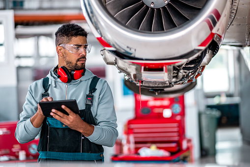 Young aircraft maintenance mechanic checks all the datas using his digital tablet in front of the jet engine in the airplane hangar.