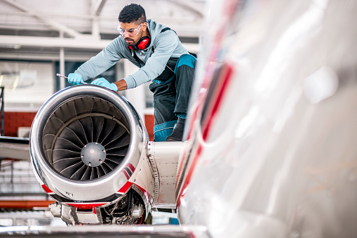 Young male mechanic seen working on top of the engine of the private jet in the hangar due to regular maintenance.