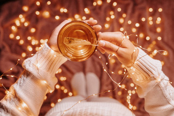 Unrecognizable young woman in cozy woolen socks and pullover and christmas lights holding a tea glass