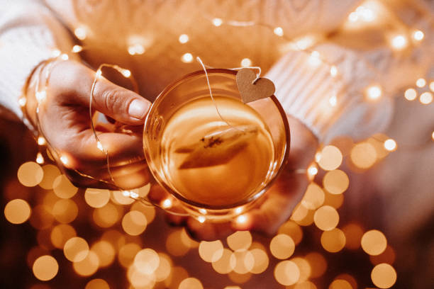 Young female hands holding a tea glass with a tea bag with a heart shape and christmas lights