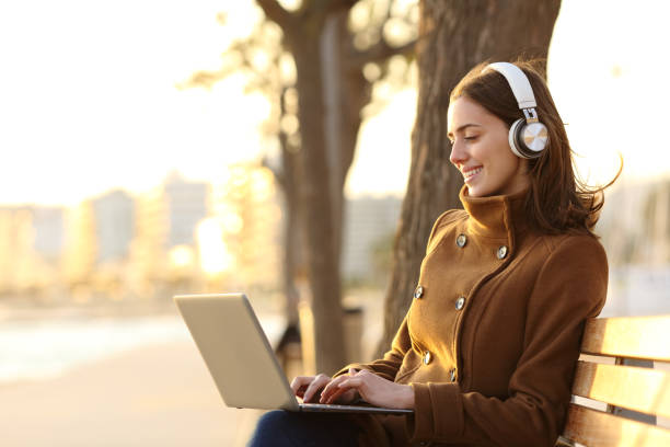 Woman wearing headphones using laptop in winter on a bench Woman wearing headphones using laptop in winter on a bench teenage girls dusk city urban scene stock pictures, royalty-free photos & images