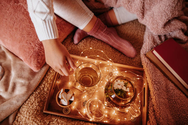 Unrecognizable young woman in cozy woolen socks on her feet and pullover with winter blankets drinking her blooming flower tea on the sofa
