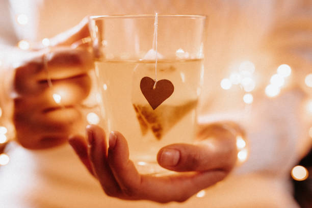 Close up of young female hands holding a tea glass with a tea bag with a heart shape and christmas lights