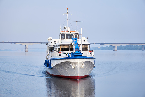 small passenger motorship on the river, front view, and a road bridge in the morning haze in the background