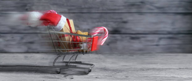 Fast small shopping cart with presents, christmas decoration, sweets and Santa Claus hat is driving on rustic wooden planks for last minute online gifts, panoramic format with copy space, motion blur rápido pequeño carrito de compras con regalos, decoración de navidad, dulces y sombrero de santa claus está conduciendo en tablones rústicos de madera para regalos en línea de última hora, formato panorámico con espacio de copia, desenfoque de movi