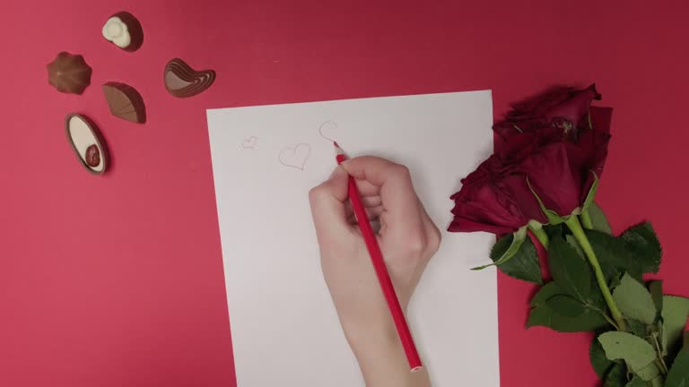 A woman draws hearts on a white sheet of paper with a red pencil. Love letter.