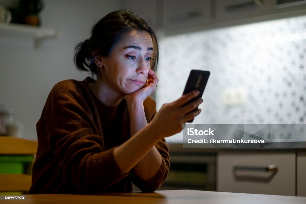 Woman looking at mobile phone screen feels upset at home at night time. 30s woman pouted lips looking at smartphone frustrated by received sms or notification, bad news reading on cell phone feels upset, waiting message from boyfriend, negative response concept. Irritation Stock Photo