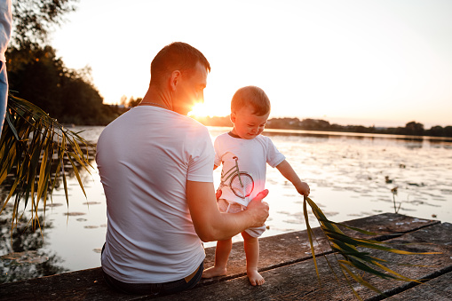 Young father and little son sitting on the wooden pier near the lake, at sunset on summer day. father's day.