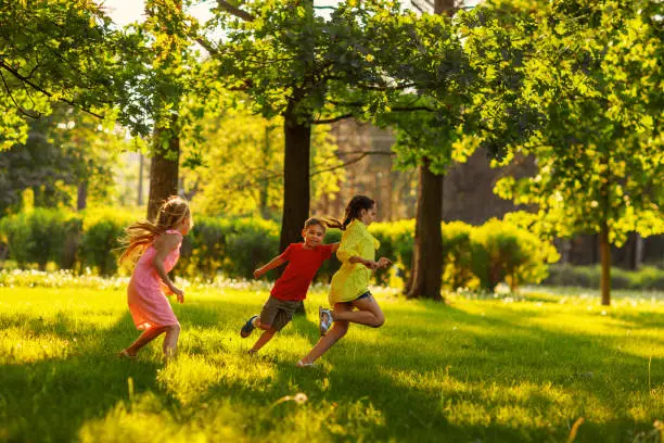 Photo of Group of three children having fun playing tag game in sunny summer park. Little friends running on green grass, chasing each other and trying to touch