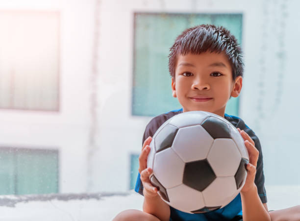 asiatische kleine fußballjunge mit lächeln hält einen fußball, während indoor zu hause zu spielen. - soccer child indoors little boys stock-fotos und bilder