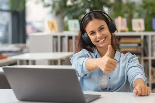 Beautiful happy young woman or student female employee sitting at table in the university library or office with laptop looking at the camera and showing thumbs up sign
