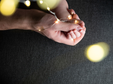 Closeup of father holding baby hand with christmas lights on gray sofa