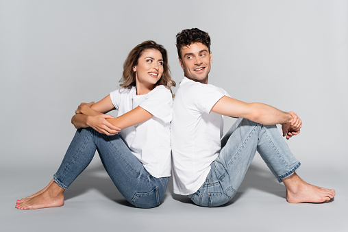 young couple in white t-shirts and jeans posing back to back isolated on grey