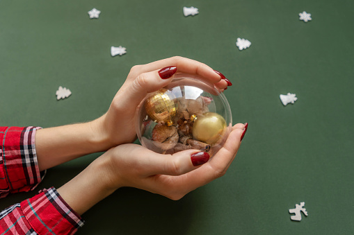 Christmas manicure. Red nails, hand with transparent ball with christmas knick-knackery inside on green background with silver baubles around.
