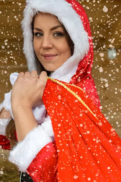 Beautiful girl Santa in red costume carrying bag of presents over her shoulder, and looking at camera with pretty smile. Close-up half-turn side portrait with snowflakes and glowing lights effect