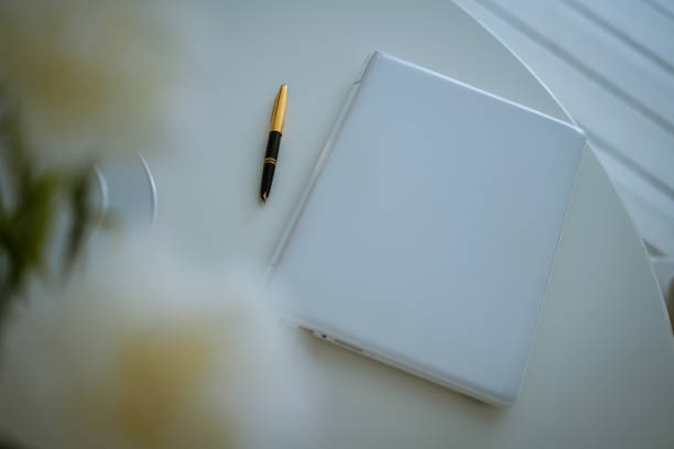Notebook and pen on a white table, flowers in a vase. Soft focus