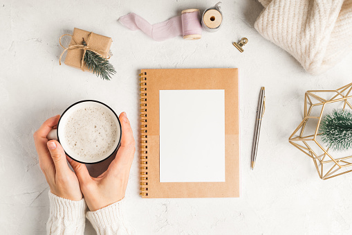 Desktop of woman hands writing in blank notepad. Flat lay of white working table background with cup of coffee and Christmas decoration. Top view, mock up greeting card, craft Notebook and pen.