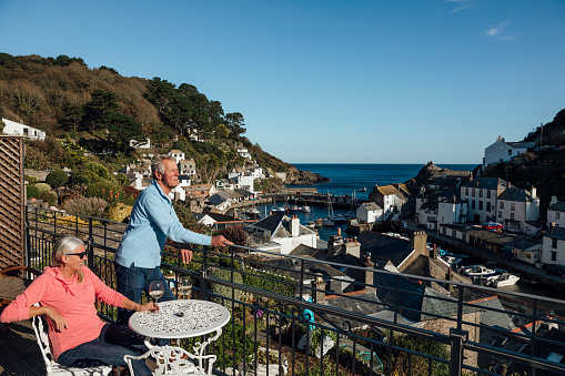A wide shot of a Caucasian, senior couple relaxing on a balcony looking over Polperro, Cornwall. The man is looking out at the view whilst his wife is enjoying a glass of wine.