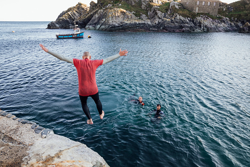 A shot of a Caucasian, senior man jumping into a harbour to join his friends at Polperro, Cornwall. He is wearing a wetsuit.