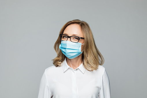 Portrait of confident mature woman wearing white shirt, N95 face mask and eyeglasses looking at camera. Studio shot, grey background.