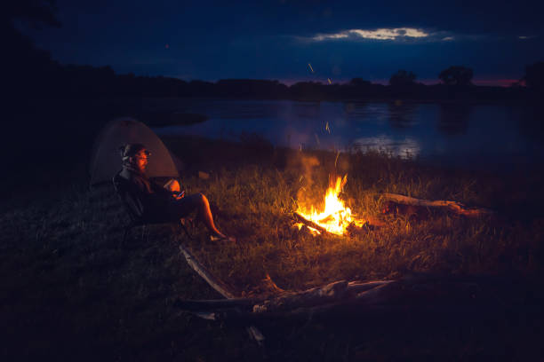 Young bearded man and sitting next to a campfire at night. A tent and river are visible in the background.