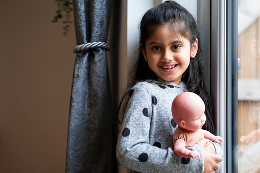 A young girl standing at the window and wearing casual clothing. She is holding a doll and looking and smiling at the camera.