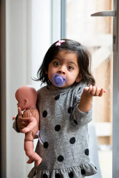 A toddler girl standing at the window and wearing pyjamas with a dummy in her mouth. She is holding a doll, looking at the camera and shrugging her shoulders.