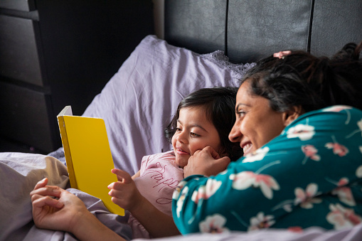 A mid adult woman and her daughter lying down in bed and reading a book together. It is the morning and they are both wearing pyjamas.