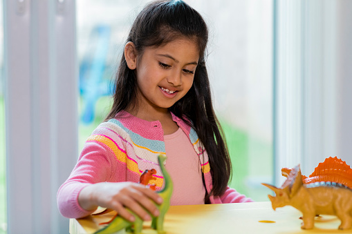 A young girl playing with her toy dinosaurs in her living room and wearing casual clothing.