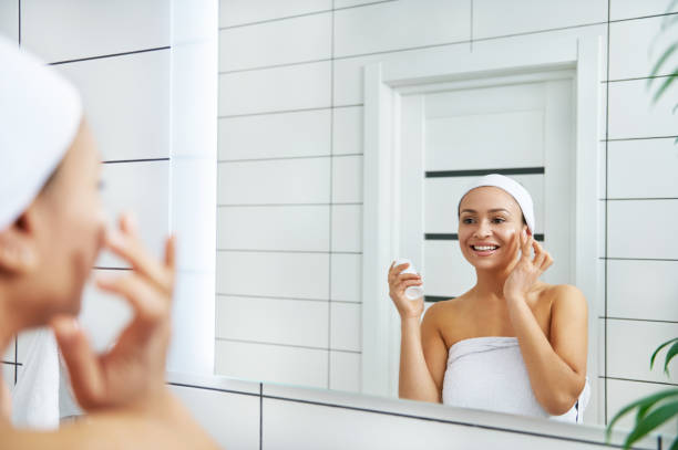 istock Woman applying cream to face with finger while looking at her reflection in bathroom mirror 1290060781
