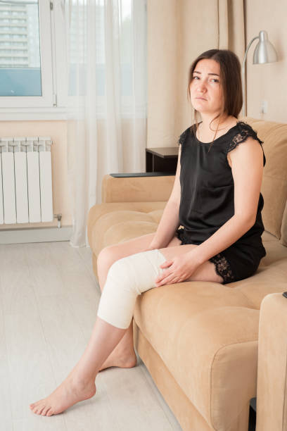 istock Sad barefoot lady with white elastic bandage on her knee sitting on sofa at home side view 1290063347