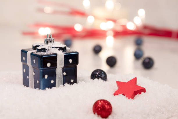 Red wooden star, blue gift box with white dots and white bow, blue and red glitter Christmas balls, snow base, soft focus background, red branches with fairy lights. Home decoration for holiday season estrella de madera roja, caja de regalo azul con puntos blancos y lazo blanco, bolas de navidad de brillo azul y rojo, base de nieve, fondo de enfoque suave, ramas rojas con luces de hadas. decoración del hogar para la temporada de vacaciones