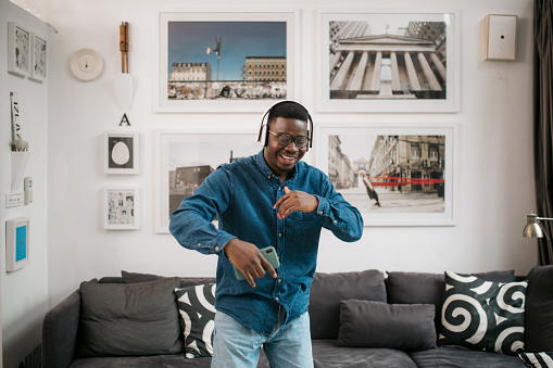 Cheerful African American young man having fun at home alone, having headphones on, listening to music using his smart phone and dancing